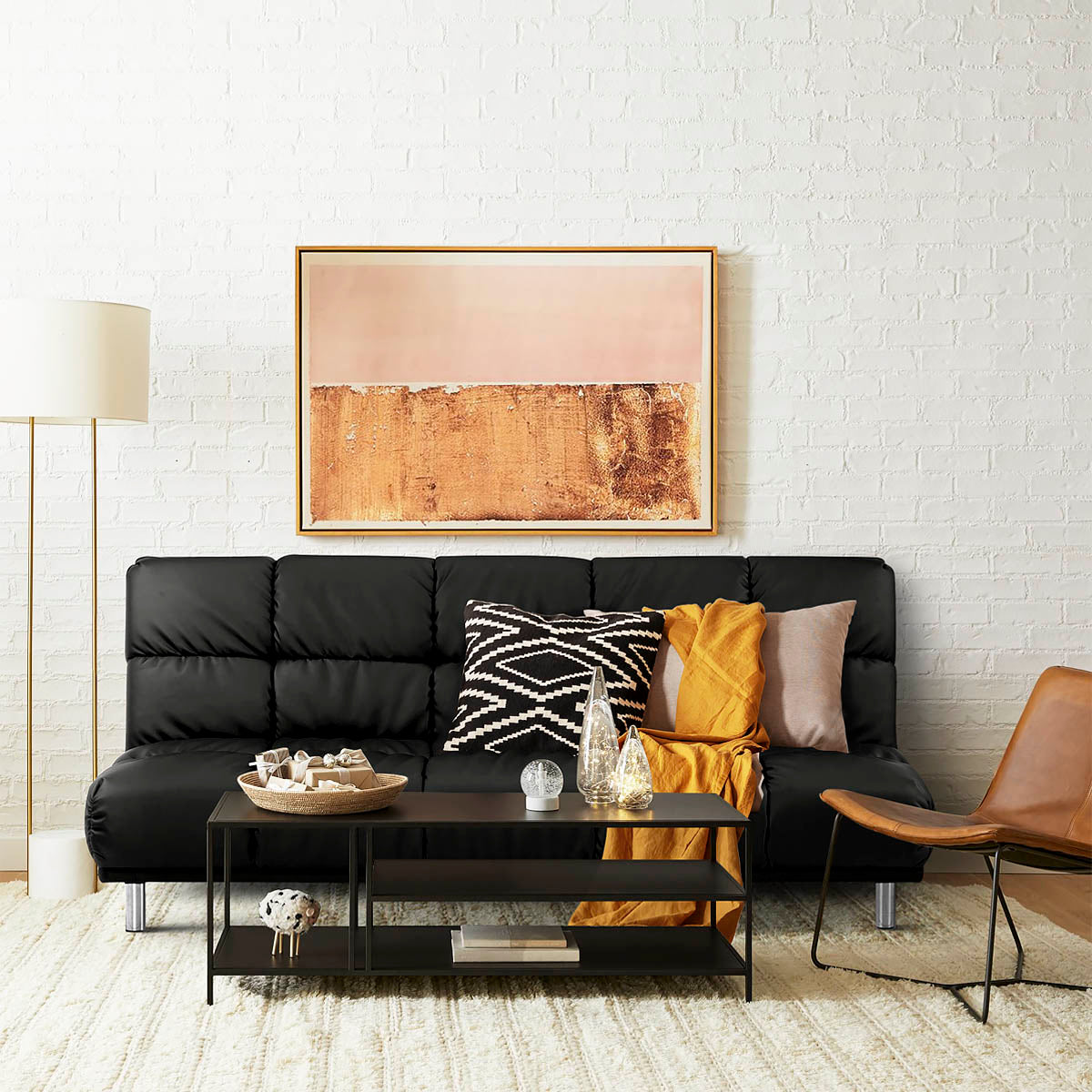 Canapé-lit clic-clac 3 places en simili cuir noir dans un salon contemporain, décoré de coussins graphiques et d’un plaid ocre, avec table basse noire, lampe sur pied et mur en briques blanches, ambiance chaleureuse et urbaine.